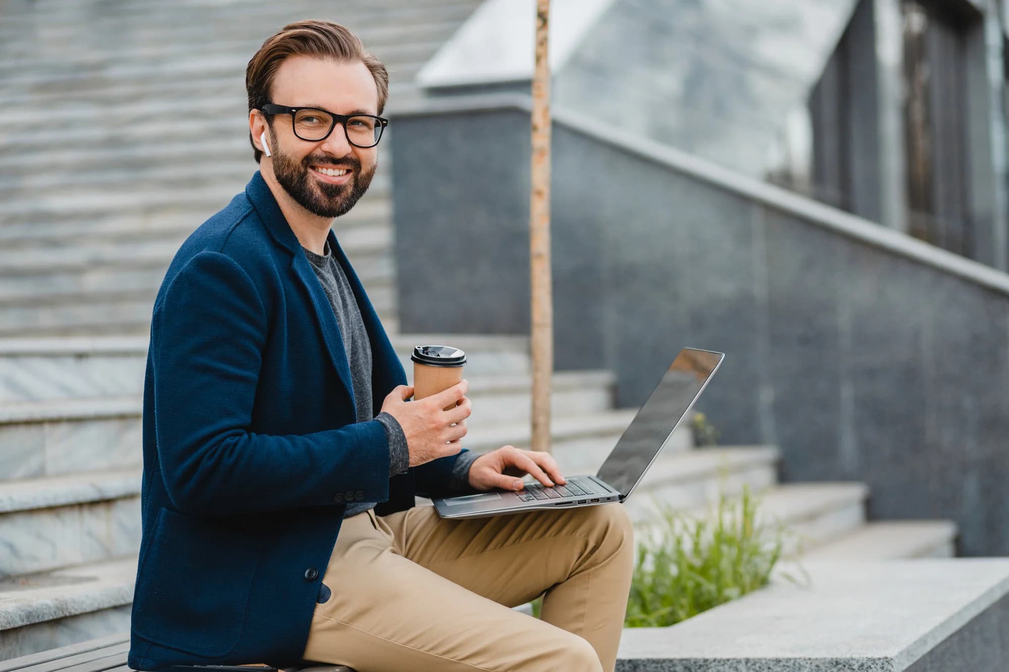 A man smiling while holding a coffee cup and working on his laptop outdoors on a stone staircase