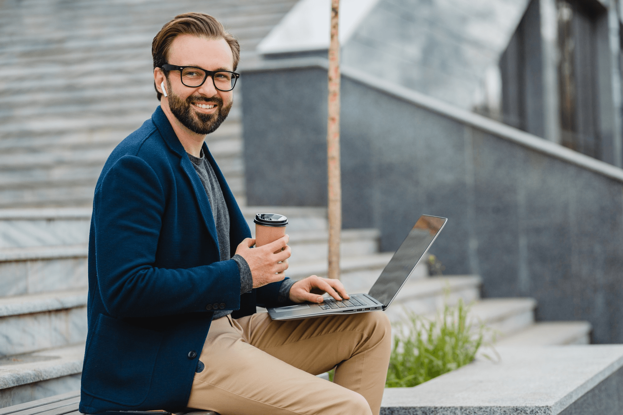 A man smiling while holding a coffee cup and working on his laptop outdoors on a stone staircase