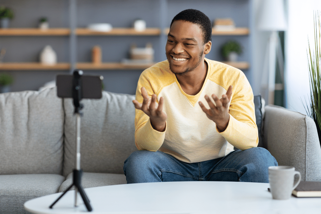 A man is sitting on a couch across from his cell phone stand. He is talking to an online audience and live streaming a webinar from his cell phone.